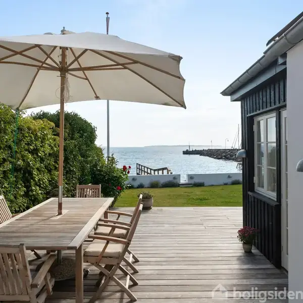 Terrassebord af træ med stole står under en stor parasol. Det hele er placeret på en træterrasse ved siden af et hus. I baggrunden ses en have og havudsigt.