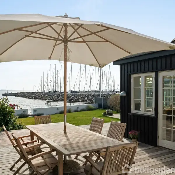 Stor udendørs parasol over træbord med seks stole på træterrasse. I baggrunden ses en marina med sejlbåde og vand mod en klar himmel.