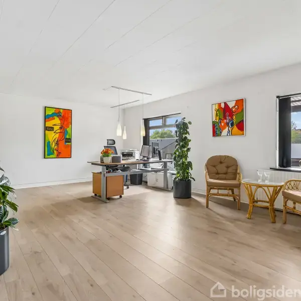 Kontorområde med to stole og et bord ved et vindue. Farverige malerier hænger på væggene. I centrum står et skrivebord med computer og planter. Lys træparketgulv.