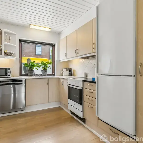 Et moderne køkken med træskabe, hvidevarer som ovn og køleskab, og en bordplade med mikrobølgeovn og planter. Vindue giver udsyn til en bygning udenfor. Trægulv og hvid loft.
