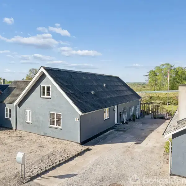 Et gråt hus med sort tag står på en åben grund ved siden af et andet hus. Der er en gårdsplads med grus og få træer i baggrunden.