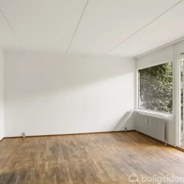 Et køkken med grå skabe og hvide vægge har trægulv. Stort vinduesparti til venstre med udsigt til have. Lang radiator løber langs højre væg.