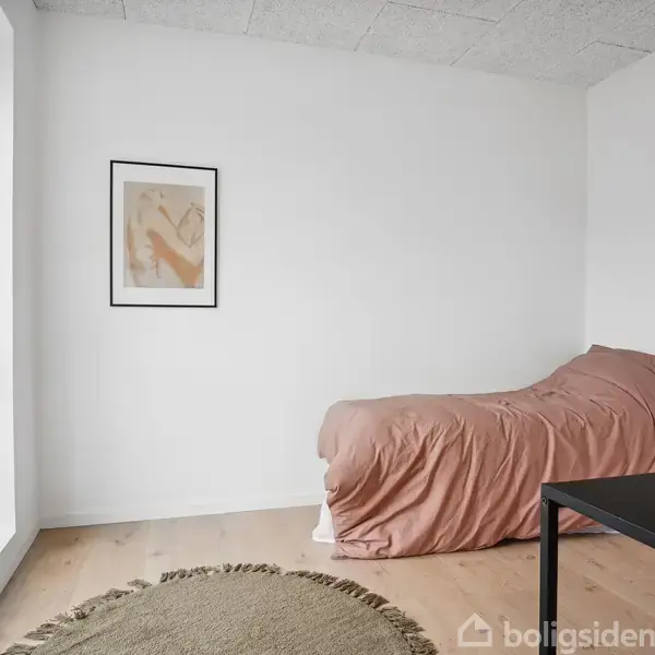 En enkeltseng med rosa sengetæppe står ved en hvid væg, dekoreret med et billede. Et rundt tæppe ligger på gulvet ved siden af et bord i en lys stue.