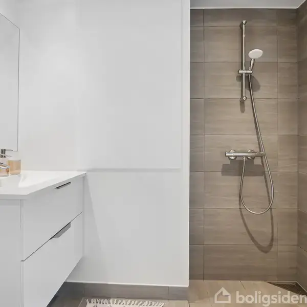 Et moderne badeværelse indeholder en væghængt bruser med brune fliser og en hvid vask med skuffer. Badeværelset har lyse vægge og en enkeltstående vask med spejl.