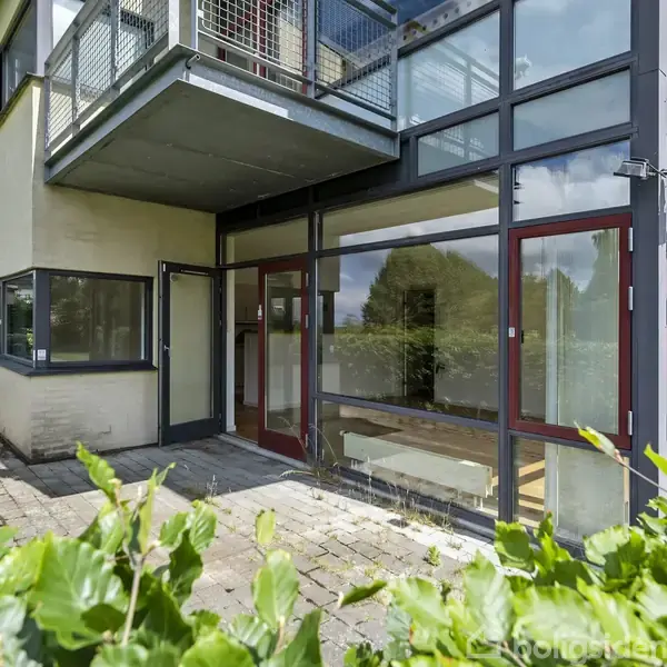 Et moderne hus med store vinduer og altan ses udefra. En flisebelagt terrasse omgives af grønne buske. Bygningen har en blanding af glas og mursten i konstruktionen.