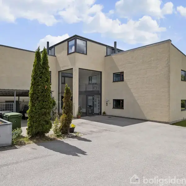 En stor, moderne bygning står stille med flere vinduer og flade taglinjer. Foran er der en asfalteret indkørsel omgivet af græsplæne og dekorative buske.