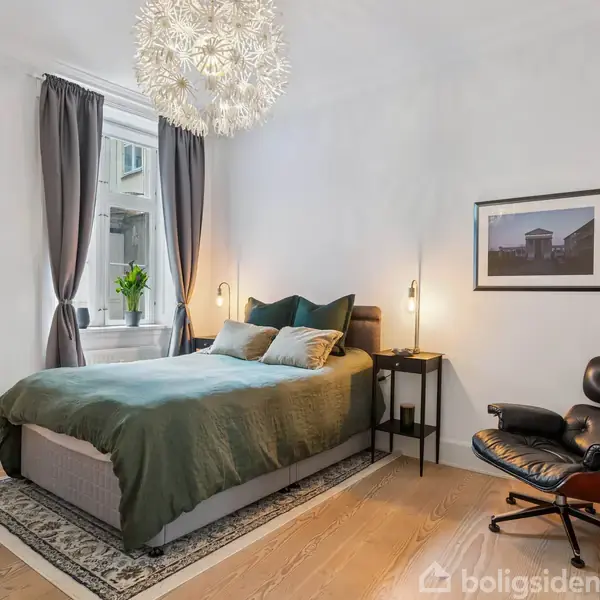 Soveværelse med en dobbeltseng med grønt sengetæppe, stillet op ad en hvid væg med billeder. En lænestol står ved siden af. En stor, lysende loftslampe hænger ned i rummet.