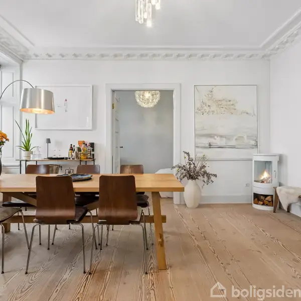 Et spisebord af træ med fire stole. En vase med blomster står på bordet. Rummet har lyse trægulve, hvidmalede vægge, og en pejs i hjørnet.