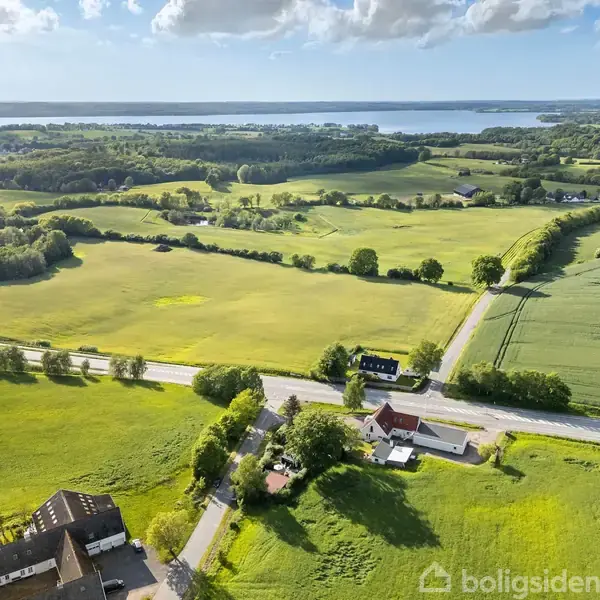 Landskab med grønne marker og spredte træer strækker sig ud mod en fjord i horisonten. Et par landeveje krydser området, omgivet af enkelte bygninger.