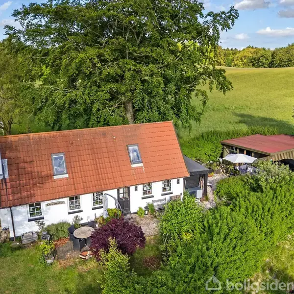 Et hvidt hus med rødt tag står omgivet af frodig grøn have, store træer og en åben mark i baggrunden under en blå himmel.