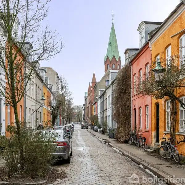 Farverige rækkehuse flankerer en brostensbelagt gade, hvor biler er parkeret langs siden, med en kirke med grønt spir i baggrunden. Træer og cykler ses langs fortovene.