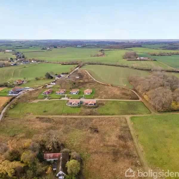 Huse står stille i en spredt klynge på åbne marker omgivet af grønne områder og skove. Rolige landeveje forbinder dem til en fjern horisont med landsbrugsmarker.