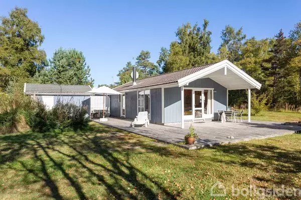 Et lyseblåt sommerhus med hvide detaljer står på en træterrasse. To stole og et bord er på terrassen. Omgivet af grønne træer og græsplæne under en klar blå himmel.
