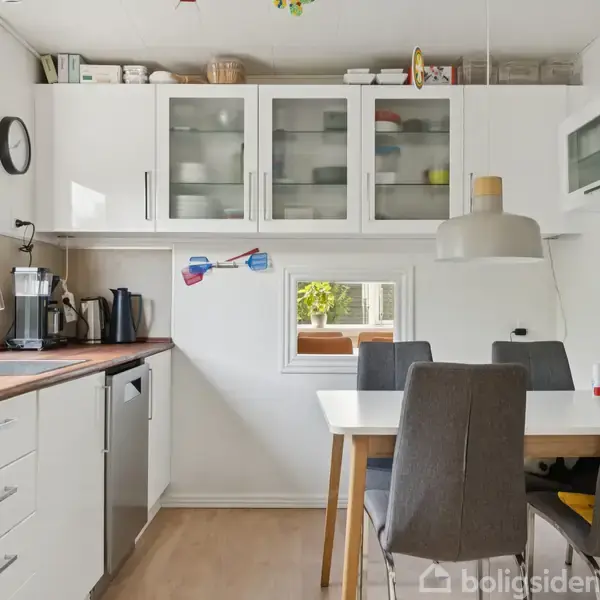 Et lyst køkken med hvide skabe og glaslåger over en træbordplade. En kaffemaskine står på bordet. Et spisebord med grå stole er placeret mod en væg med et lille vindue.