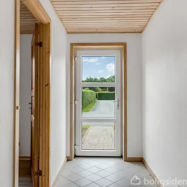 Dør i glas-og-træ-ramme står åben mod en havevej. Døre til venstre leder til andre rum. Lyse klinker dækker gulvet, og væggene er hvide.