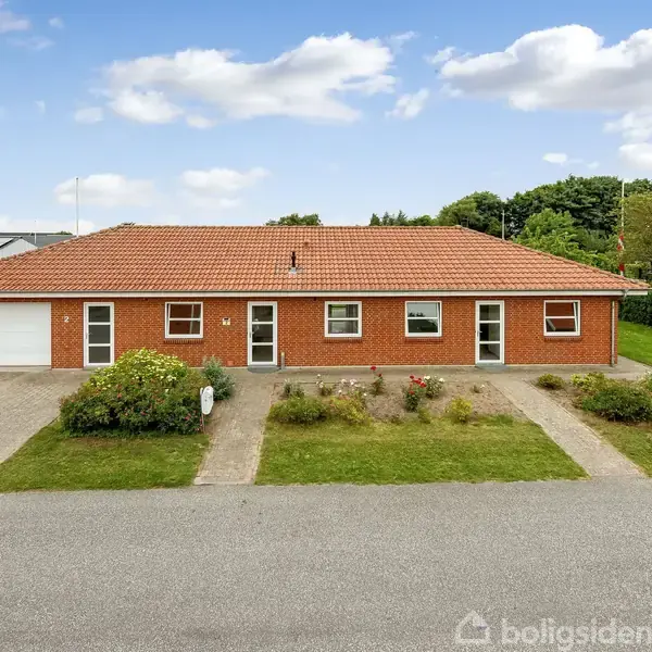 Et rødt murstenshus med tre indgangspartier og garage. Beliggende i et boligområde med velplejede græsplæner og buske foran. Skyfri himmel med enkelte skyer i baggrunden.