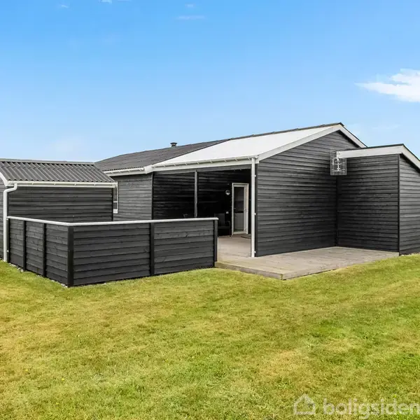 Et sortmalet sommerhus står roligt med moderne design, omgivet af en græsplæne og en rød bygning i baggrunden under en klar blå himmel.