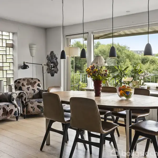 En spisebord står i et lyst rum med træstole omkring det. En glasvase med blomster og en frugtskål pryder bordet. To lænestole står ved siden af, omgivet af store vinduer og grøn udsigt.