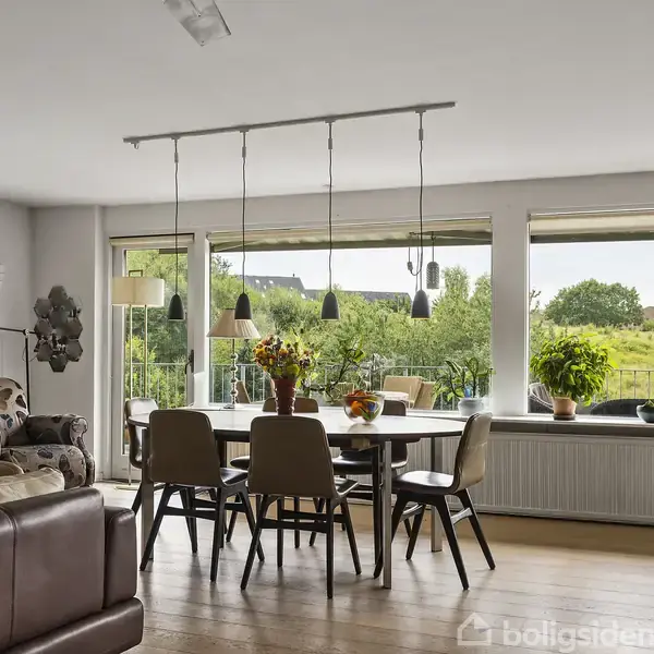 Spisestue med træbord omgivet af stole, oplyst af hængelamper. Store vinduer viser udsigt over grøn have. Sofaer og planter tilføjer hygge i rummet.