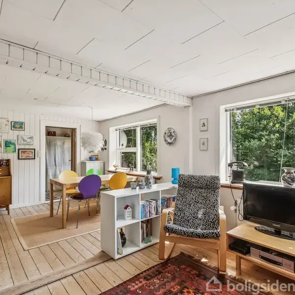 En stue med et TV på et lavt bord, en mønstret stol og bogreol i forgrunden, leder til en spiseplads med farverige stole omkring et bord, omgivet af store vinduer med udsigt til frodig natur.