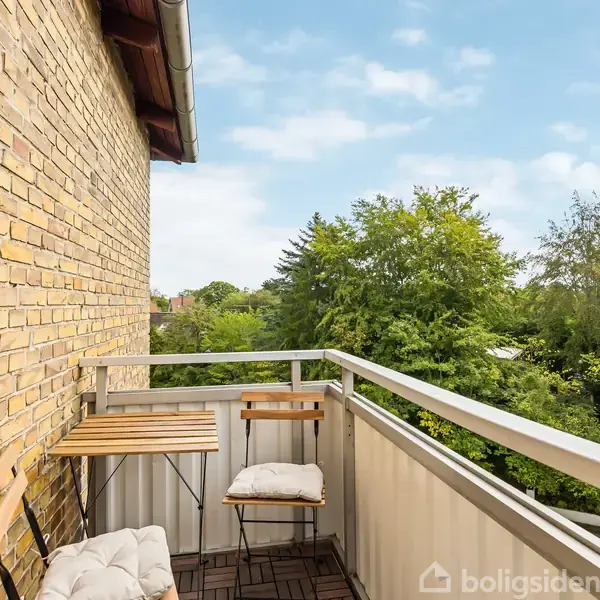 Balkon med et lille cafébord og to stole, placeret mod en husmur af gule mursten. Udsigt til grønne træer under en delvis skyet himmel.