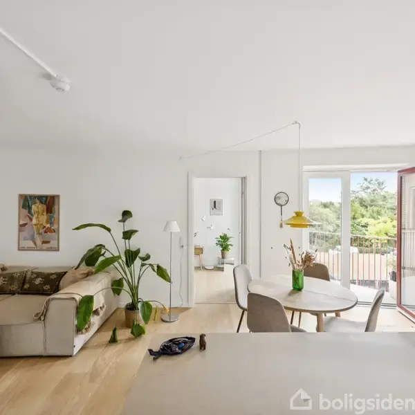 Stue med en beige sofa og grønne planter, beliggende i et lyst rum. Spisebordet er dækket og står nær en åbentstående altandør med udsigt til grønne omgivelser.