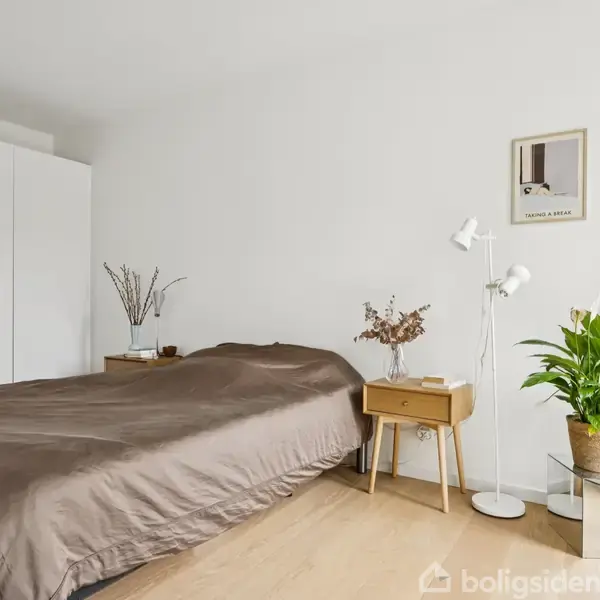 Soveværelse med dobbeltseng dækket af brun dyne, omgivet af to natborde. En plante og lampe står ved siden af. I baggrunden ses et skab og en minimalistisk vægkunst.