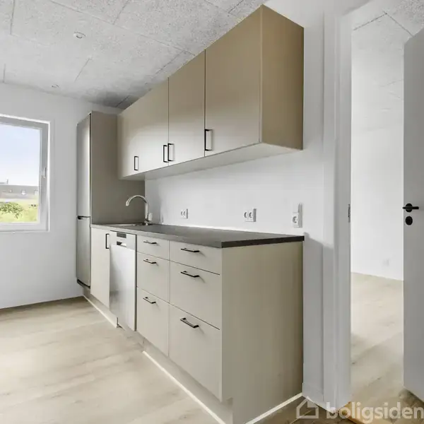 Køkken med beige skabe og skuffer langs væggen, en vask under et vindue, og en åben dør til højre. Flisegulv og hvidmalede vægge skaber et lyst rum.