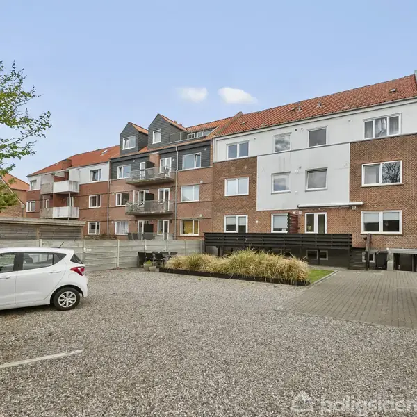 Lejlighedskompleks med røde teglstenstag og altaner står stille i en gårdsplads. En hvid bil er parkeret på en grusplads, omgivet af grønne buske og hegn.
