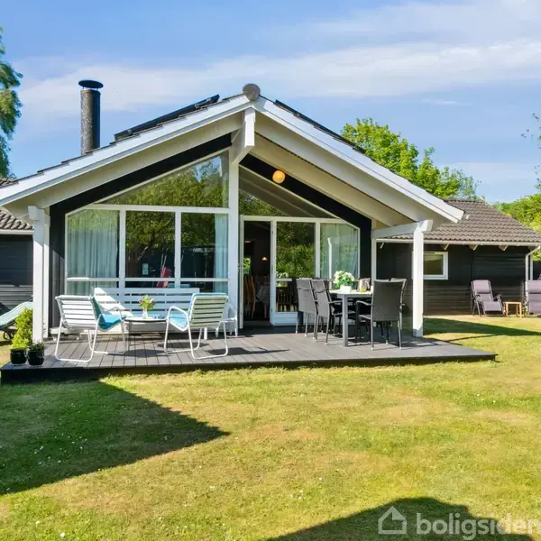 Sommerhus med sort træbeklædning står på en stor græsplæne. Terrassen har hvide havemøbler. Omgivet af grønne træer og klar blå himmel.