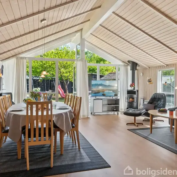 Spisebord med træstole står dækket i en hyggelig stue; store vinduer lukker lys ind fra haven, mens sofaer og brændeovn skaber en varm atmosfære med træbeklædte vægge og loft.