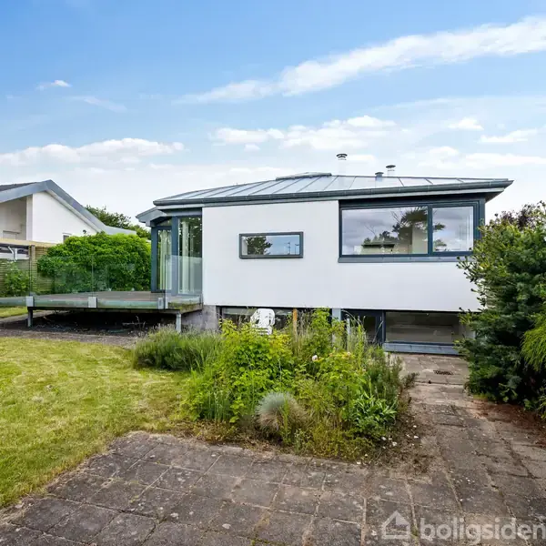 Hus står stille ved grøn have; moderne, hvid facade med store vinduer. Terrasse på venstre side. Naboer ses til venstre under blå himmel.