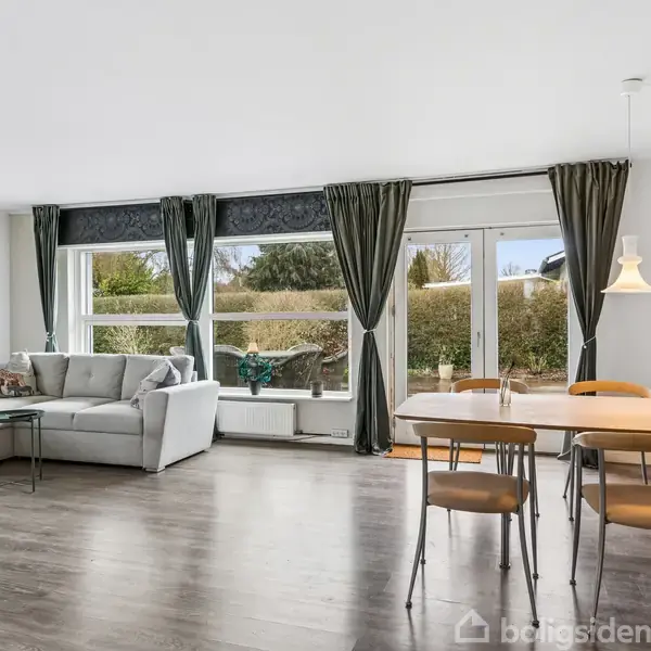 Sofa står i en lys stue ved store vinduer med udsigt til haven, flankeret af grønne gardiner. Ved siden af er et spisebord med stole og hængelampe.