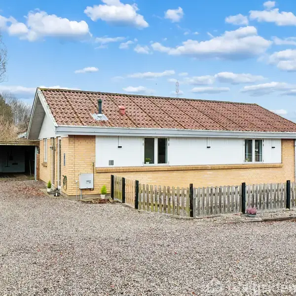 Et enfamilieshus med rødt tegltag og gule mursten står stille ved siden af en grusindkørsel. En garage ses til venstre og en hegn omkranser en have.