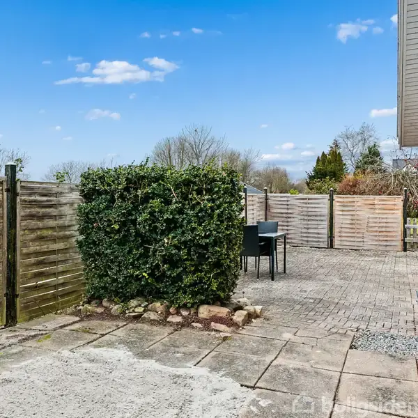 Bord og stol står stille på en flisebelagt terrasse omgivet af en træhegn og en grøn hæk; et hus med murstensvæg er på højre side under en blå himmel.