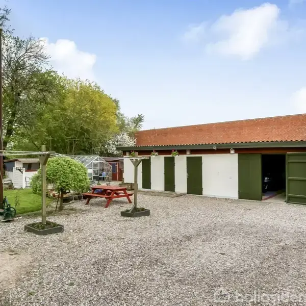 En grusbelagt gårdsplads med en træbænk og et tørrestativ. I baggrunden ses en bygning med grønne døre og rødt tag samt en have med træer og drivhus.