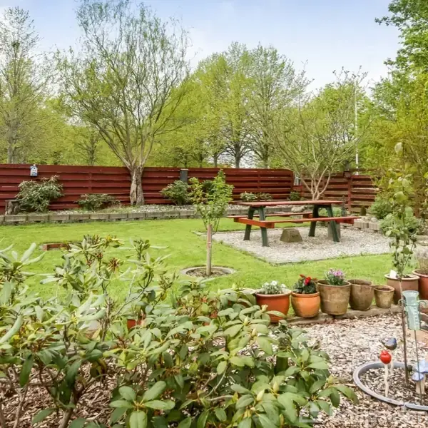 Bord-bænkesæt står i en grøn have omgivet af blomsterkrukker og buske. Rød træhegn indrammer området, og høje træer ses i baggrunden. Skilt i højre forgrund.