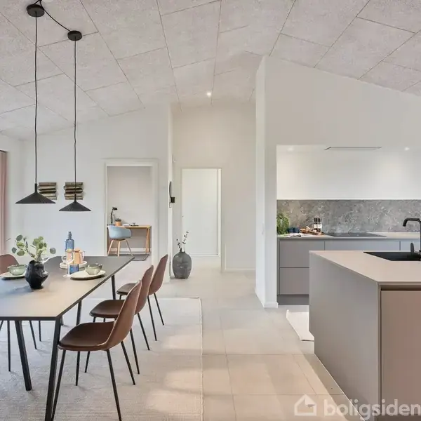 Spisebord med brune stole står i et moderne køkken-alrum. Der hænger sorte lamper over bordet, og til højre ses en køkkenø. Store vinduer giver udsigt til en grøn have.