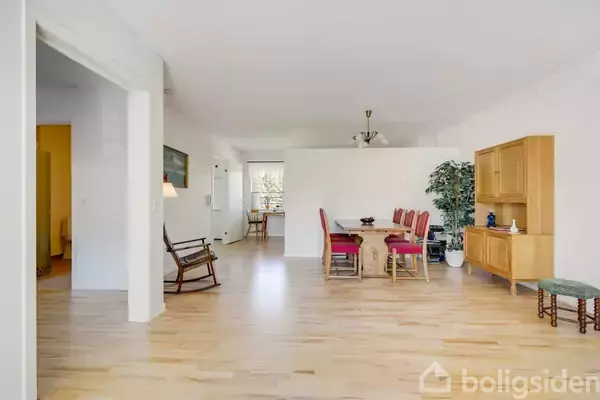 En spisestue med et træbord og seks røde stole står centralt. Til højre ses et træskab og en plante. Væggene er hvide, og gulvet er lyst trægulv.