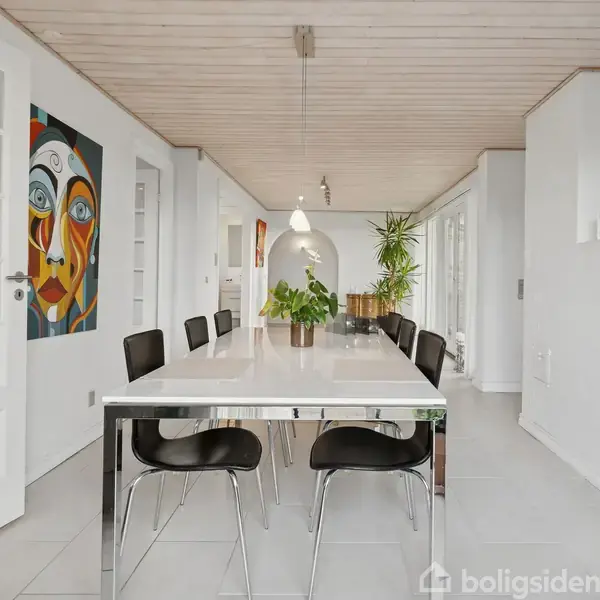 Langt spisebord med sorte stole står i et lyst rum med hvide vægge og træloft. Et farverigt ansigtsmaleri hænger ved en dør. Planter er nærmest vinduet.