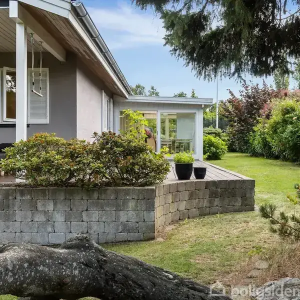Hus med terrasse og havemøbler står på en lav murstensplatform. En frodig have omkranser terrassen, og en trægren ligger på græsset ved siden af.
