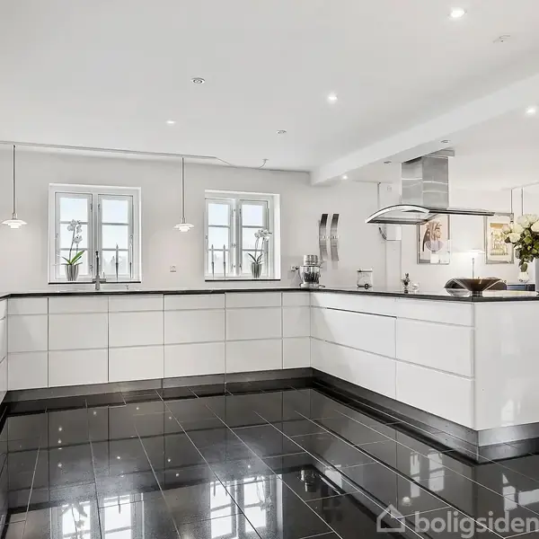 Et moderne køkken med hvide skabe og sorte fliser strækker sig i en vinkel, mens tre vinduer giver naturligt lys. I baggrunden er der en stue med sofa og dekorationer.