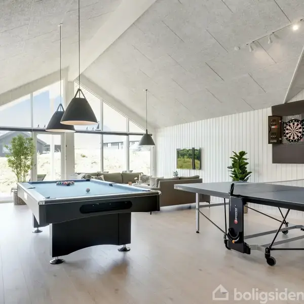 Poolbord og bordtennisbord i aktivitetsrum med højt loft og store vinduer. Et dartskab hænger på den hvide væg. Grønne planter tilfører rummet liv.