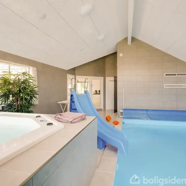 Indendørs swimmingpool med rutsjebane i et lyst rum. I forgrunden ses et boblebad og et håndklæde på kanten. Planter og lyse vægge skaber en afslappende atmosfære.