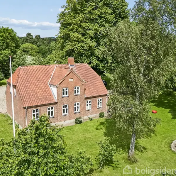 Rødt murstenshus med rødt tegltag står midt i en grøn, skovrig have. En flagstang og en trampolin ses foran huset. En stor træterrasse spænder over baghaven.