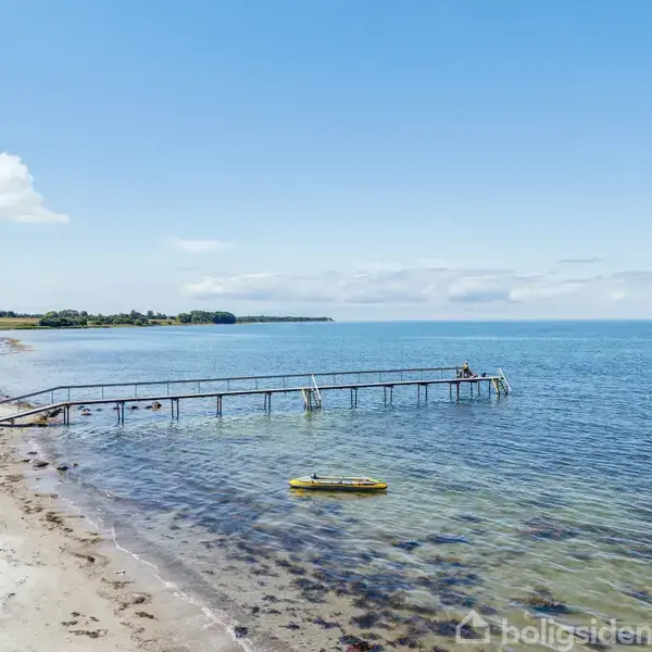 Mole strækker sig ud i det rolige, blå hav fra en sandstrand. En gul oppustelig båd flyder tæt på kysten. Spredte skyer dekorerer den klare himmel.