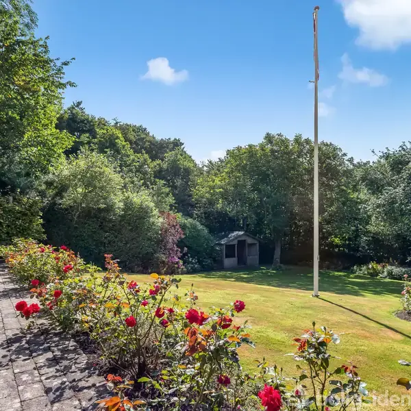 Terrasse med bord og stole står stille, omgivet af have med blomsterbede. En græsplæne pryder midten, flagstang står oprejst, og skur står i baggrunden nær træer og buske.