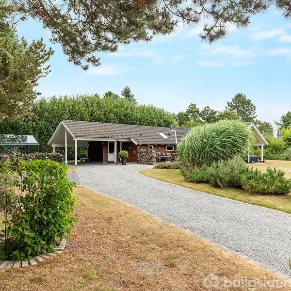 Hus med fladt tag og carport i en frodig have. Grusvej fører op til huset, som er omgivet af høje træer og planter, hvilket skaber en fredfyldt atmosfære.
