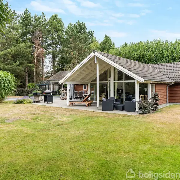 Et sommerhus med store vinduer står stille i en velplejet have omgivet af høje træer. En terrasse med havemøbler og grill er placeret foran huset i den grønne omgivelser.