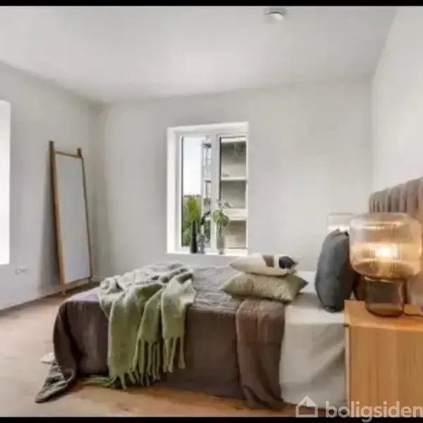 En seng står i et lyst soveværelse med tæpper og puder. Et natbord med en lampe er ved siden af sengen. Vinduet viser udsigt til bygninger udenfor.